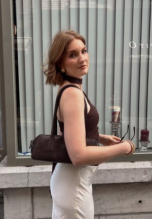 Bolso de hombro marrón hecho de cuero con cierre de cremallera, forma rectangular y asas cortas. Llevado por una persona con un top marrón y una falda blanca.