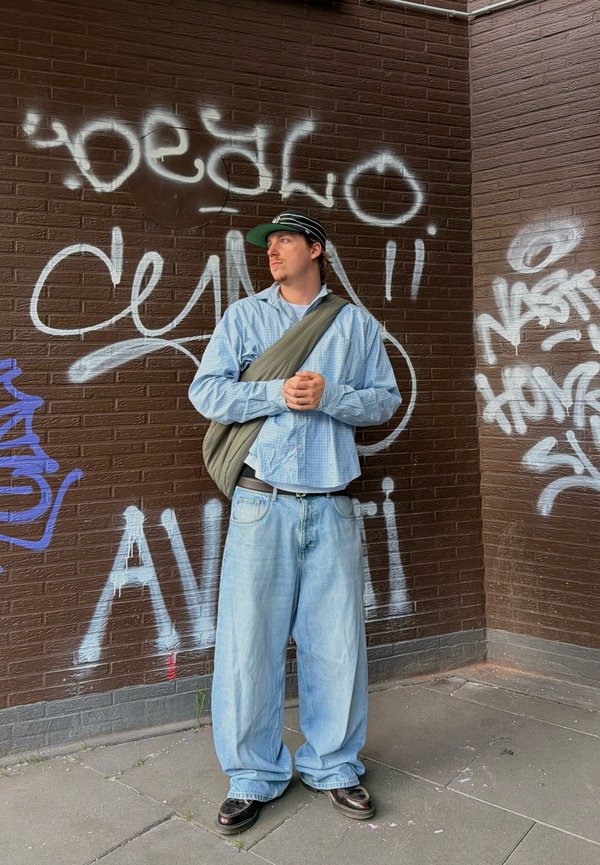 Jeune homme en jean large, chemise bleue et casquette, debout les mains jointes contre un mur en briques brunes couvert de graffitis sur un trottoir.