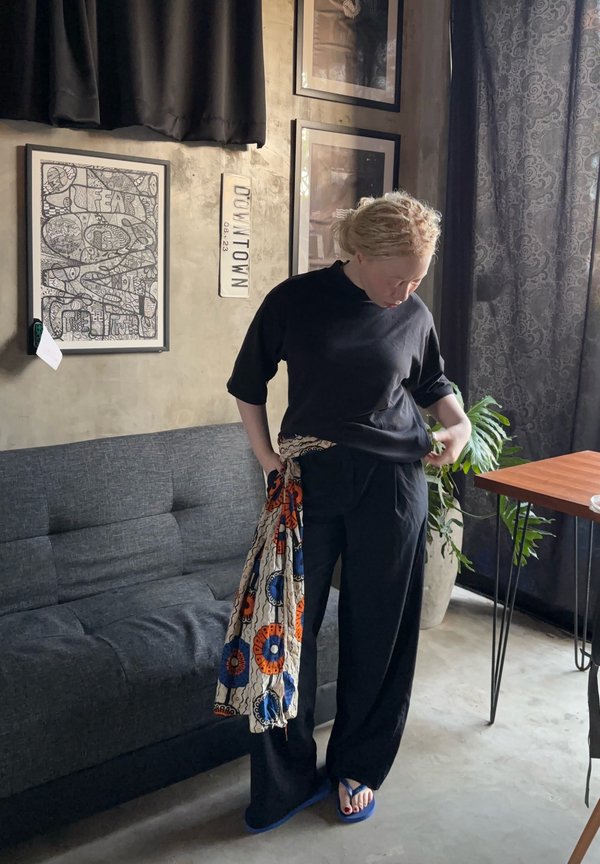 Persoană cu păr deschis la culoare, îmbrăcată în negru, stă în interior lângă o canapea gri, ținând o țesătură colorată cu modele, cu artă încadrată și o plantă în fundal.