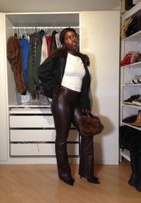 Fekete bombázó dzseki szőrös gallérral, fehér crop top, barna műbőr harangnadrág és barna szőrös kézitáska. Fából készült padló háttér.
