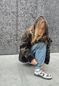 Nike Dunk αθλητικά παπούτσια σε γκρι και λευκό με καφέ λεπτομέρεια, με ανώτερο μέρος από υφαντό δέρμα και ύφασμα, τοποθετημένα σε γκρι πλακάκι.