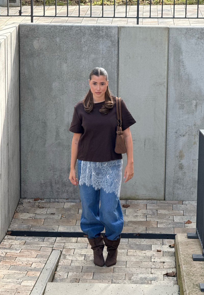 Jeune femme portant un haut marron, un pantalon bleu avec une superposition en dentelle, et des bottes marron, montant des marches en pierre en plein air à côté de murs en béton.