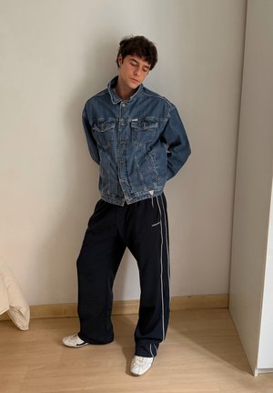 Chaqueta vaquera con bolsillos delanteros, botones y un cuello estructurado, llevada sobre pantalones deportivos oscuros con franjas blancas laterales y zapatos blancos casuales.