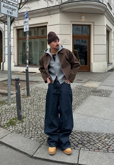 Chaqueta de pana marrón sobre un hoodie gris, combinada con unos jeans de corte ancho y botas de color marrón claro. De pie contra una calle de adoquines texturizados.