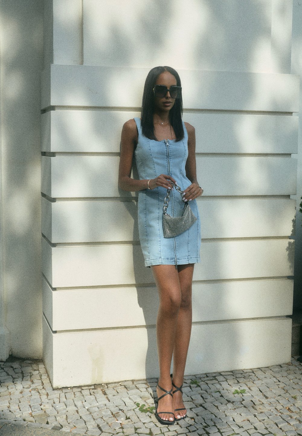 Robe en denim bleu clair avec fermeture éclair sur le devant, fines bretelles et coupe ajustée, associée à un mini sac à main métallique et des sandales noires à lanières.