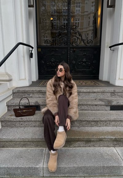 Mujer con cabello largo y castaño, vestida con un abrigo de piel color canela y pantalones marrones, sentada en escalones de piedra, ajustándose los zapatos cómodos beige, con un bolso a su lado.