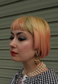 Kort bob-frisyre med gradientfarger fra gul til oransje. Tilbehør inkluderer store gulløreringer og flere lag med perlekjeder.