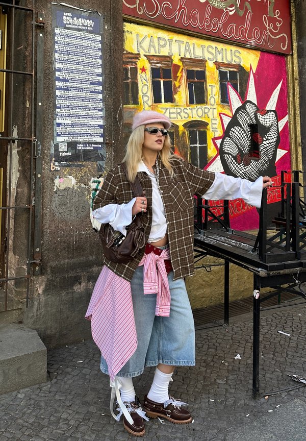 Ung kvinde med solbriller, lyserød baret, ternet skjorte og denim culottes står foran graffiti med løftet næve og anti-kapitalistisk budskab.