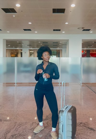 Sudadera negra con cremallera, leggings negros y zapatos color topo. Botella de agua transparente en mano. Maleta gris con ruedas al lado. Espacio interior moderno.