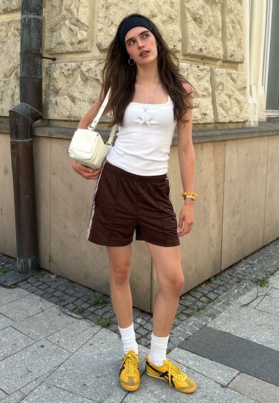 Joven mujer con pantalones cortos marrones, camiseta sin mangas blanca, zapatillas amarillas y cinta negra en la cabeza está de pie en la acera de la ciudad cerca de una pared de piedra.