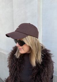 Barna baseball sapka ívelt karimával, szövetből készült. Nagy fekete napszemüveggel és sötét barna bojtós külső réteggel viselve.