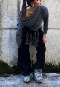 Femme avec queue de cheval se penche en avant contre un mur texturé, portant un pull gris à épaules dénudées, un pantalon noir, une chemise à carreaux nouée à la taille et des bottes argentées.