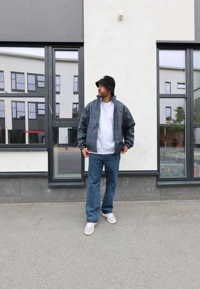 Chaqueta bomber de cuadro en verde oscuro y azul marino, camiseta gris, vaqueros azules y zapatillas gris. Hombre de pie frente a un edificio con grandes ventanas.