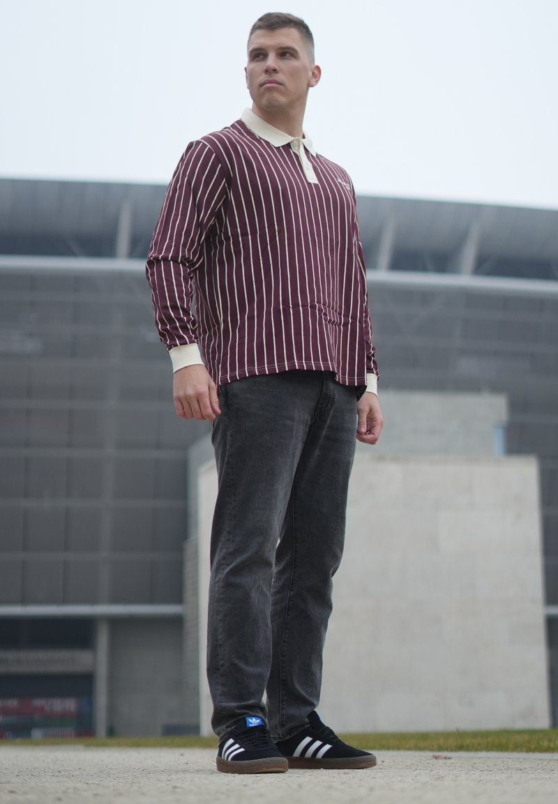 Polo a maniche lunghe a righe bordeaux e bianche, jeans grigi, sneakers nere con strisce bianche, in piedi all'aperto davanti a uno sfondo moderno.