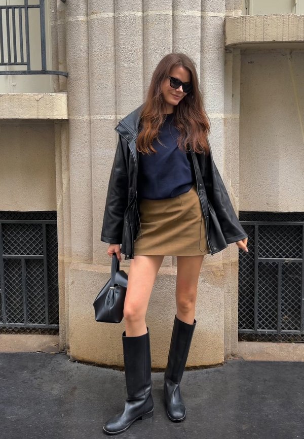 Mujer con gafas de sol, chaqueta de cuero negra, falda marrón y botas hasta la rodilla posa frente a un edificio de piedra, sosteniendo un bolso negro.