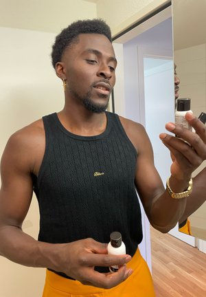 Hombre con una camiseta sin mangas negra texturizada con un acento dorado examina dos botellas pequeñas, una blanca con una tapa oscura y una oscura. Suelo de madera al fondo.