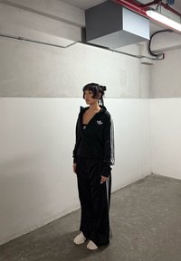 Asmuo su pintomis plaukų kasomis, apsirengęs juodu "Adidas" treniruočių kostiumu ir baltais sportiniais batais, stovi tuščioje pramoninėje patalpoje su pilkais ir baltais sienomis.