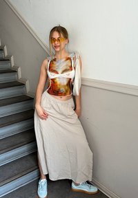 Weißes Crop-Top mit abstraktem orange- und grau-muster, beiger Maxi-Rock und hellblauen Schuhen. Die Accessoires umfassen gelbe Sonnenbrillen.