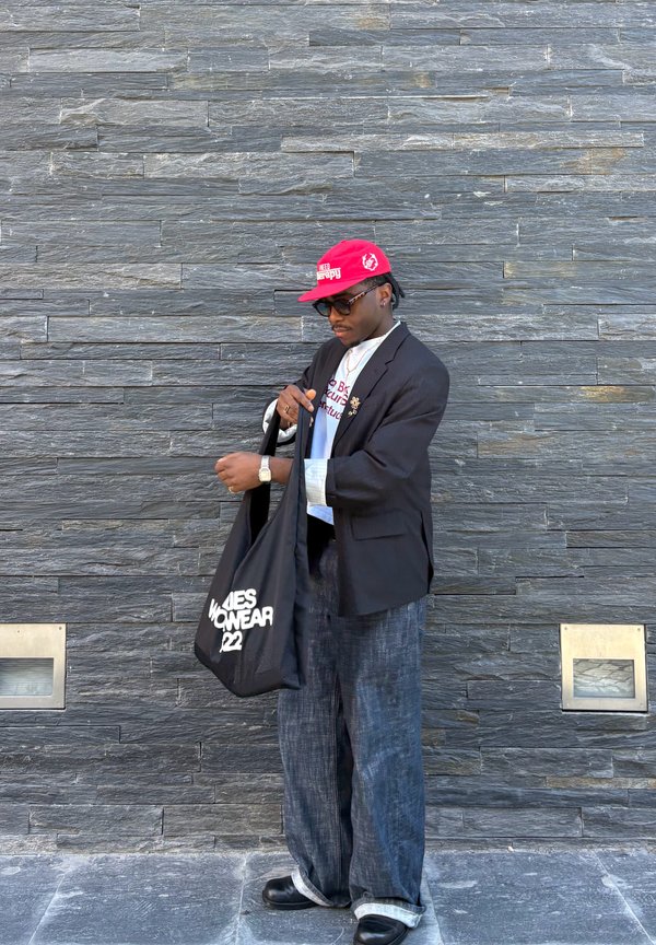 Άνδρας που φοράει κόκκινο καπέλο, γυαλιά ηλίου, μαύρο σακάκι, φαρδιά τζιν και μαύρα παπούτσια, κρατώντας μια μαύρη τσάντα ώμου με λευκό κείμενο μπροστά σε έναν πέτρινο τοίχο.