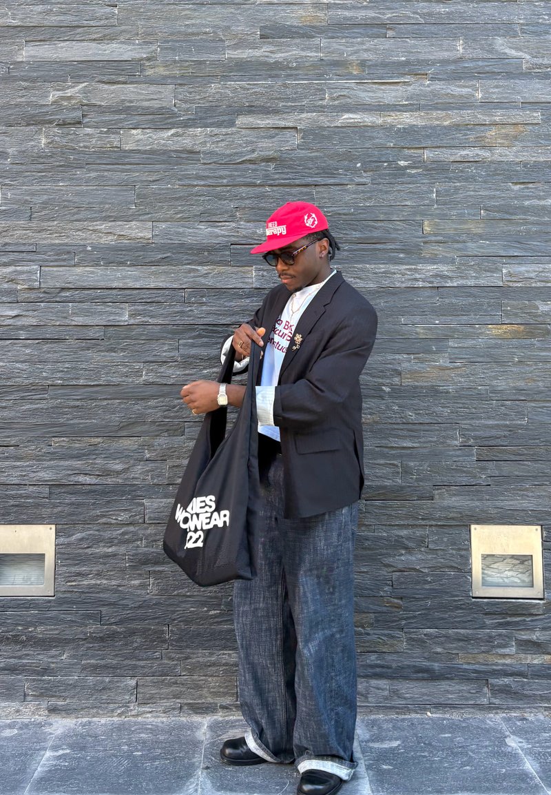 Homme portant une casquette rouge, des lunettes de soleil, un blazer noir, un jean ample et des chaussures noires, tenant un sac fourre-tout noir avec un texte blanc devant un mur en pierre.