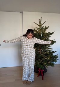 Bézs és fehér kockás pizsamaszett fa mintákkal, gombos felsővel, hosszú ujjú, bő szabású nadrággal, bézs papuccsal egy díszített szobában.