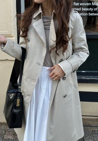 Trencho beige de doble botonadura con epaulettes en los hombros, grandes botones y un corte relajado, combinado con una falda blanca y un bolso negro.