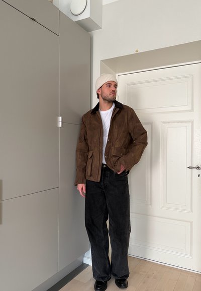 Hombre con chaqueta marrón, gorro blanco, camisa blanca y pantalones anchos negros está de pie en el interior junto a un armario gris y una puerta blanca.