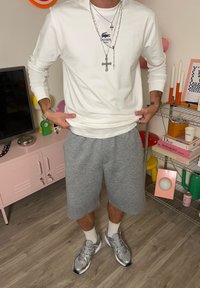 Camisa blanca de manga larga con logo, pantalones cortos grises, zapatillas deportivas grises y calcetines. Los accesorios incluyen múltiples cadenas de plata con colgantes de cruz.