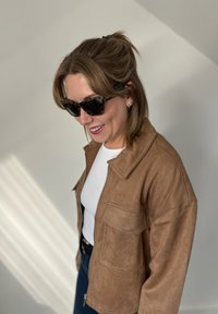 Brown suede jacket with a zip front, side pockets, and a collar. Worn over a white ribbed top with dark denim jeans. Black sunglasses worn.