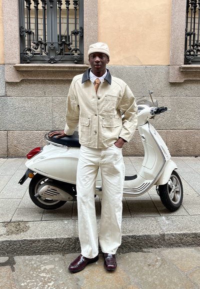 Chaqueta de lona beige con cuello gris, camisa blanca y corbata marrón. Pantalones off-white y zapatos marrones. Scooter blanco en el fondo.