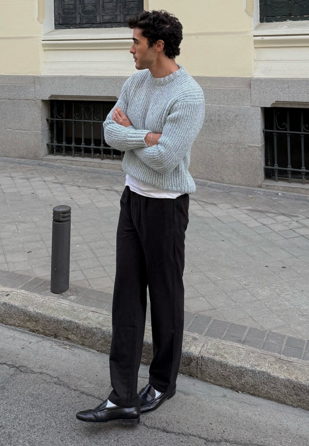 Maglione a coste azzurro chiaro, sottile camicia bianca, pantaloni ampi neri e mocassini neri. Ambiente urbano con pavimentazione in pietra.