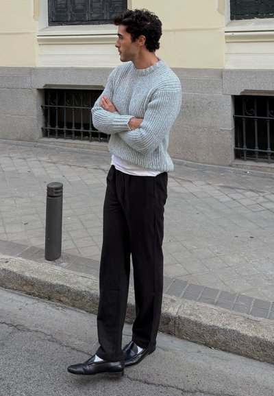 Suéter de punto acanalado azul claro, camiseta blanca debajo, pantalones anchos negros y mocasines negros. Escenario urbano con pavimento de piedra.