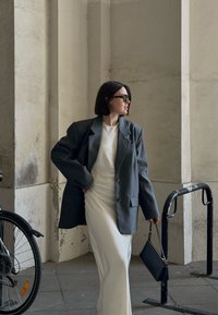 Overmaatse grijze blazer met knopen over een getailleerde witte jurk, gecombineerd met zwarte zonnebril en een kleine zwarte handtas. Stedelijke omgeving.