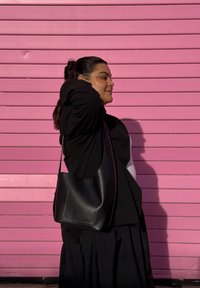 Mala de ombro em pele preta com textura suave, forma curva e alça única, segurada contra um fundo às riscas cor-de-rosa.