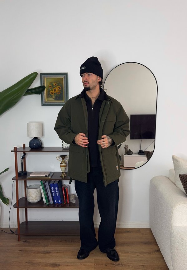 Casaco verde-oliva com múltiplos bolsos sobre uma camisa preta, combinado com jeans escuros e um gorro preto. Ambiente interno minimalista.