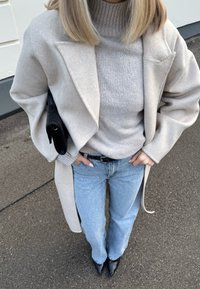 Personne portant un manteau beige et un pull à col roulé gris, un jean bleu clair, des chaussures noires pointues, et tenant un sac noir sous le bras.