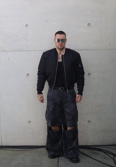 Chaqueta bomber negra sobre una camiseta sin mangas negra, combinada con pantalones cargo sueltos de patrón oscuro con grandes recortes y bolsillos. Gafas de sol plateadas.