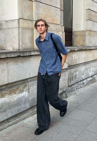 Camisa de cuadros azul de manga corta, jeans negros holgados, zapatos negros y un bolso de hombro, caminando junto a un muro de piedra en un entorno urbano.
