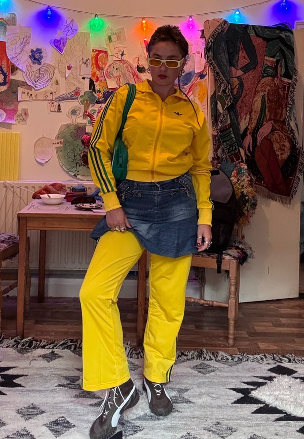 Persona con tuta gialla brillante, gonna di jeans e borsa verde, in piedi in una stanza colorata con disegni e luci sul muro.
