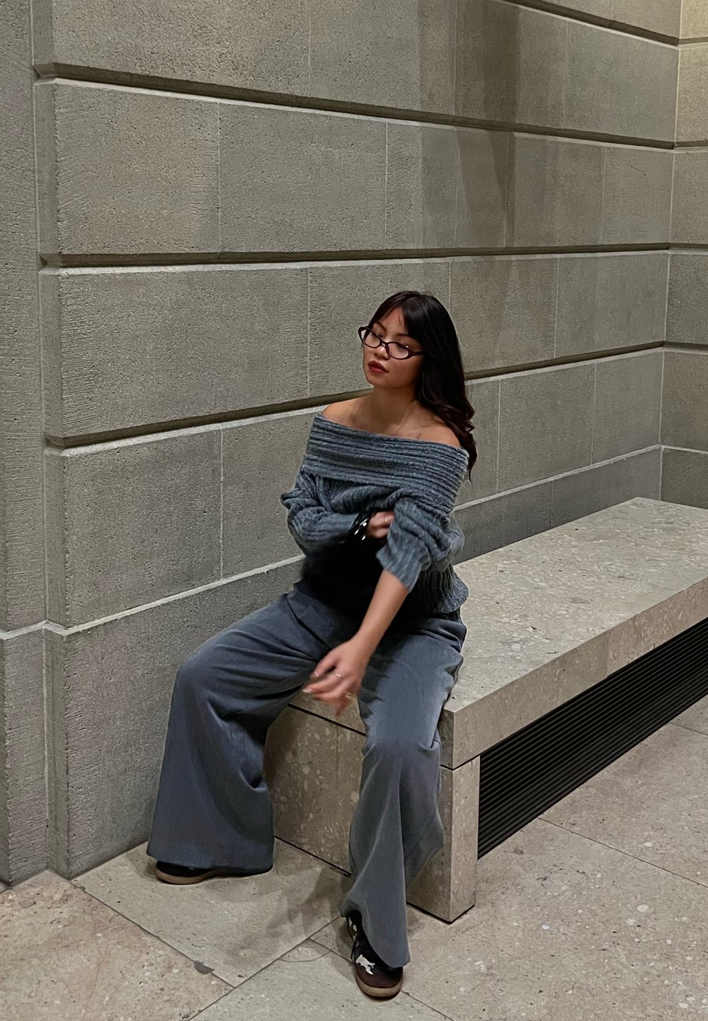 Jeune femme portant des lunettes et un pull gris à épaules dénudées assise sur un banc en pierre contre un mur carrelé à l'intérieur.