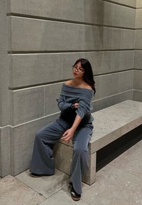 Jeune femme portant des lunettes et un pull gris à épaules dénudées assise sur un banc en pierre contre un mur carrelé à l'intérieur.