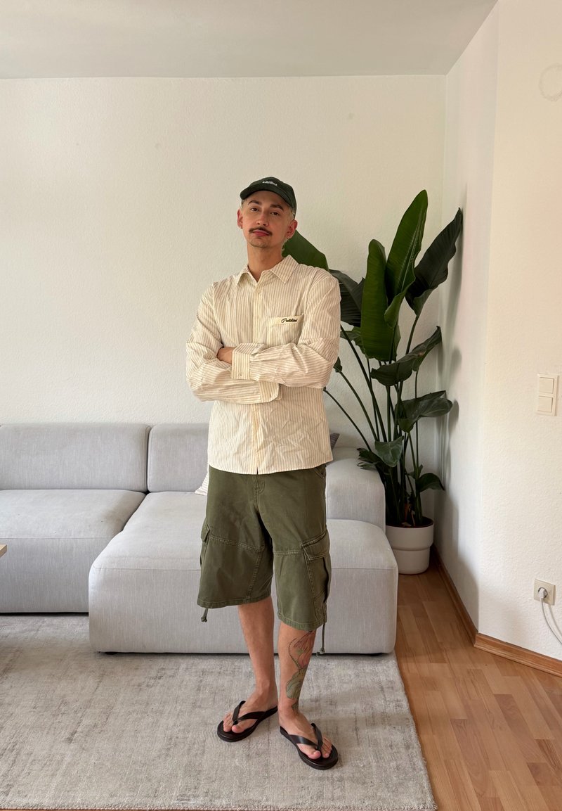 Homme debout, bras croisés, portant une casquette, une chemise à rayures, un short vert et des tongs dans un salon avec un canapé et une grande plante.