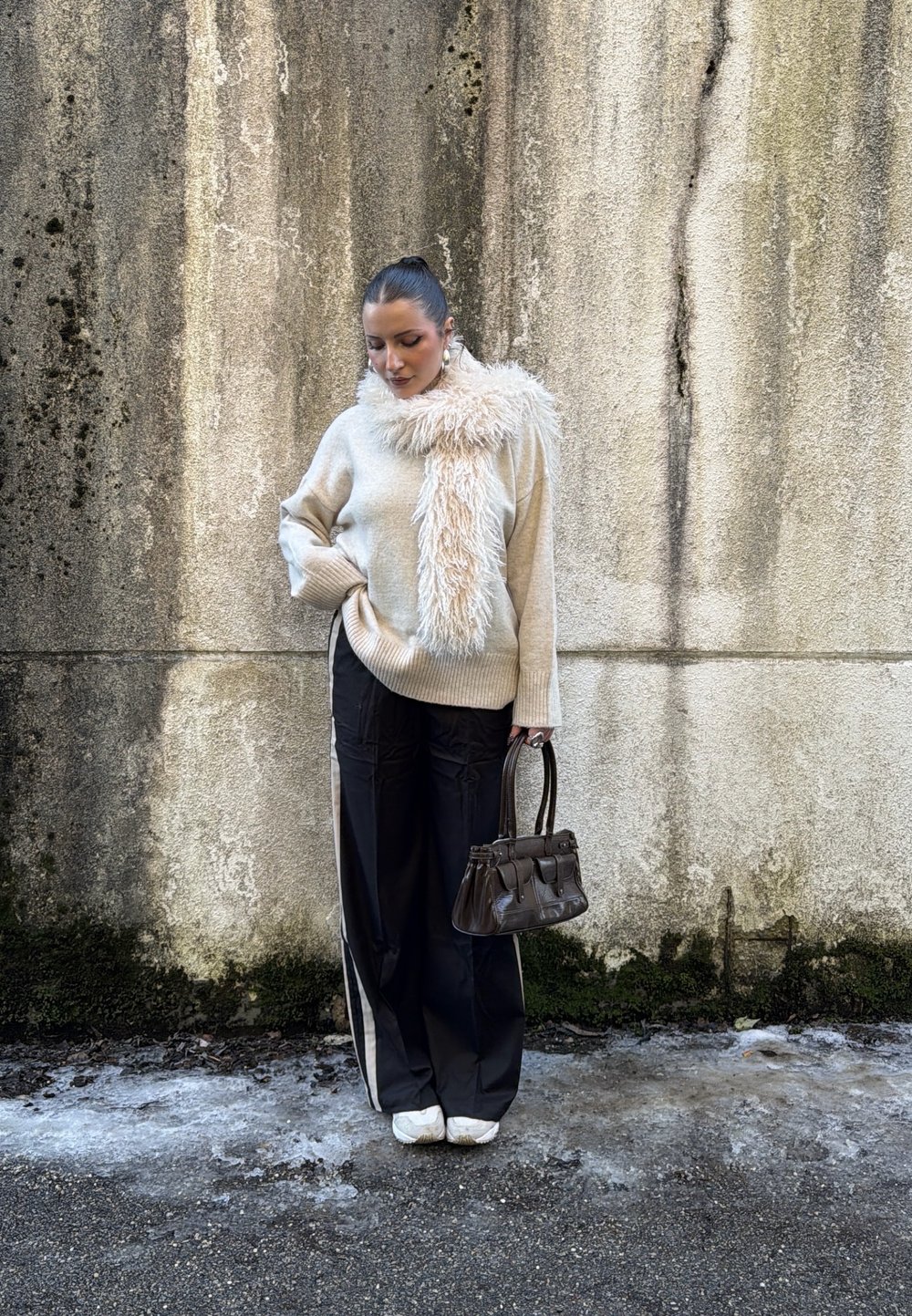 Femme portant un pull beige et une écharpe duveteuse, un pantalon large noir à rayures blanches, des baskets blanches, tenant un sac à main foncé, debout contre un mur texturé.