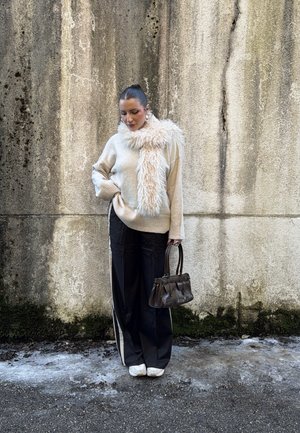 Mujer con suéter beige y bufanda esponjosa, pantalones anchos negros con rayas blancas, zapatillas blancas, sosteniendo un bolso oscuro, de pie contra una pared texturizada.