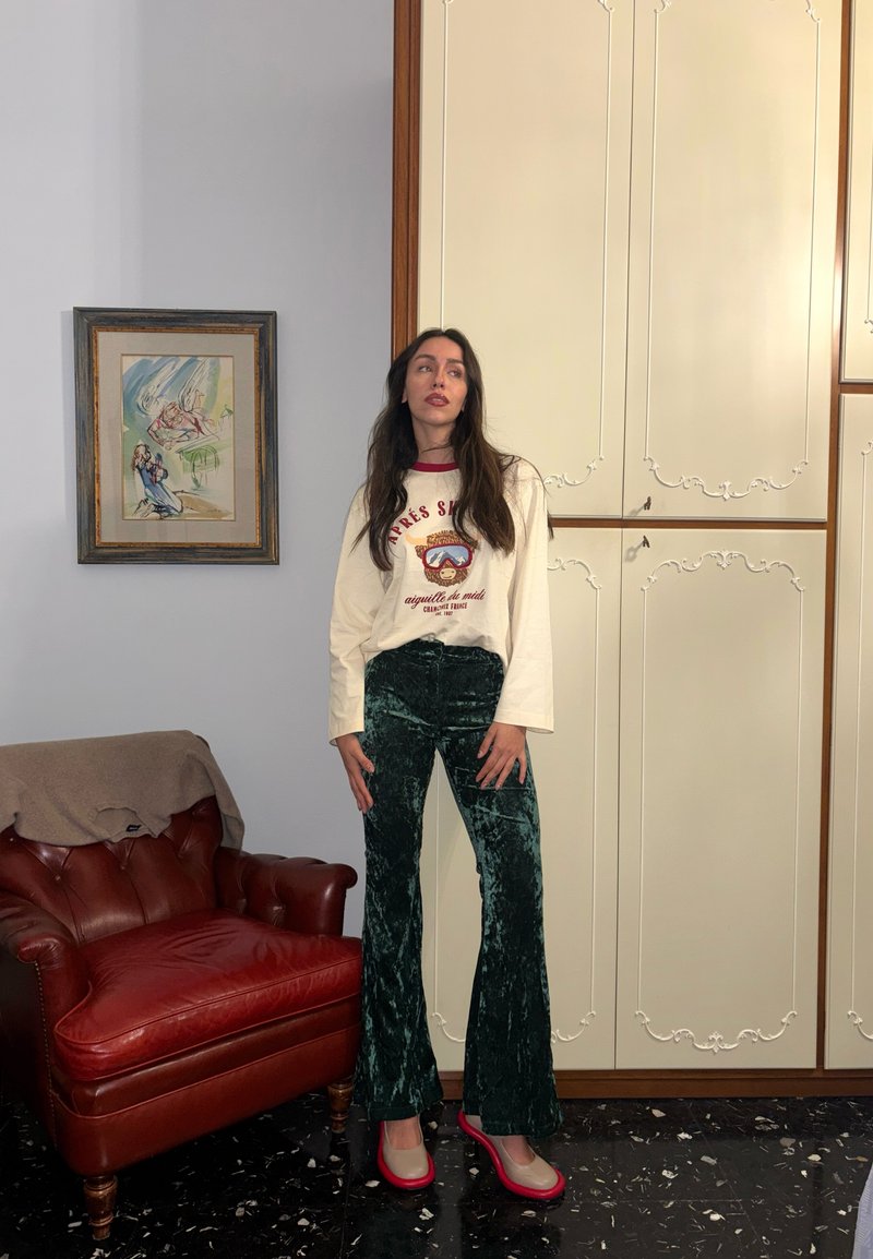 Donna in piedi all'interno vicino a armadietti color crema e un quadro incorniciato, indossa una camicia bianca a maniche lunghe, pantaloni di velluto verdi e scarpe bicolore.