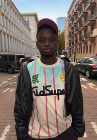 Black bomber jacket worn over a light mint shirt decorated with pink vertical stripes, featuring green and yellow logos; paired with a light pink cap.