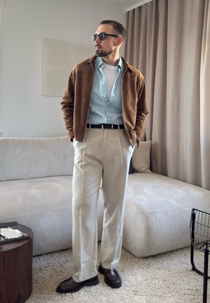 Hombre con chaqueta marrón, camisa azul claro, pantalones beige, zapatos oscuros y gafas de sol de pie en una sala de estar moderna con sofá y cortinas beige.