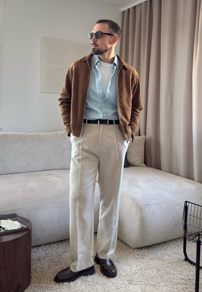 Hombre con chaqueta marrón, camisa azul claro, pantalones beige, zapatos oscuros y gafas de sol de pie en una sala de estar moderna con sofá y cortinas beige.