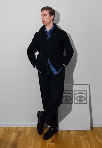 Abrigo de lana negro sobre una camisa azul, pantalones negros y zapatos lustrados. De pie contra una pared lisa con una obra de arte enmarcada que muestra ojos.