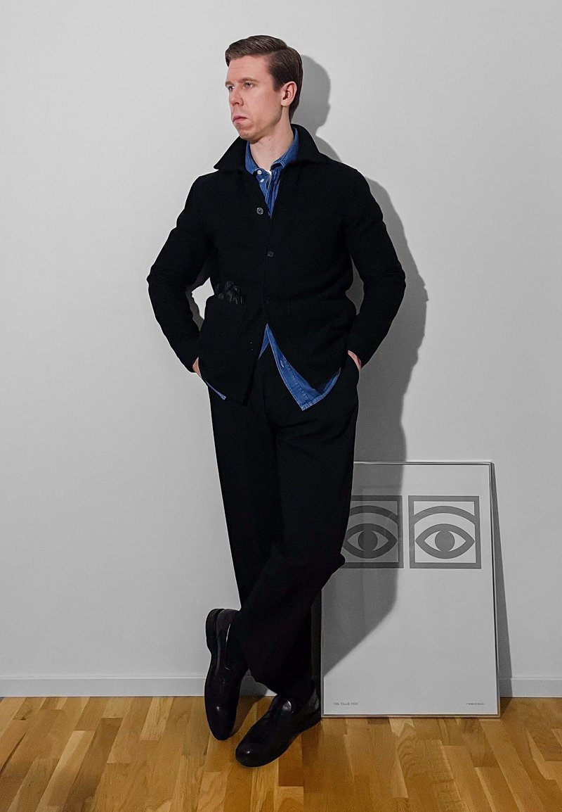 Schwarzer Wollmantel über einem blauen Hemd, schwarze Hose und polierte Schuhe. Steht vor einer schlichten Wand mit gerahmten Kunstwerken, die Augen zeigen.